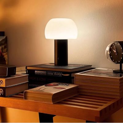 Lampe champignon noire abat-jour blanc allumé sans fil rechargeable posée sur meuble bois avec livres réveil matin