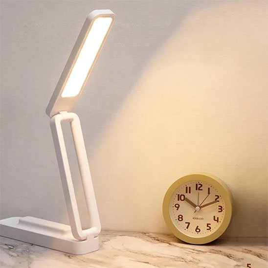 lampe de bureau LED blanche et rose pliable et polyvalente tactile et rechargeable la lampe est allumée avec une lumière tout à côté d'un réveil matin