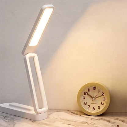 lampe de bureau LED blanche et rose pliable et polyvalente tactile et rechargeable la lampe est allumée avec une lumière tout à côté d'un réveil matin