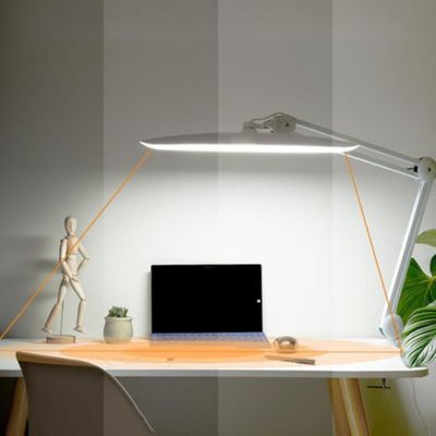 Lampe de bureau LED blanche avec bras articulé posée sur bureau blanc éclairant espace travail avec mannequin bois écran noir plante verte et chaise beige