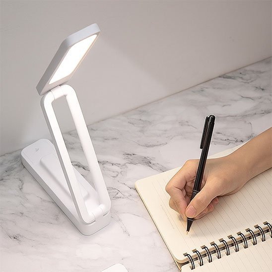 Lampe de bureau LED pliable tactile variétés d'intensité la lampe est posée sur une table pendant que l'on voit une main qui écrit profitant de la lumière