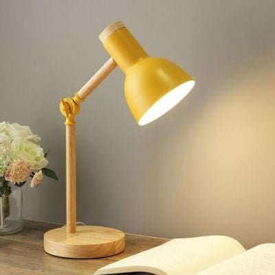 lampe de bureau jaune moutarde avec base bois et bras articulé allumée sur table de chevet avec livre ouvert et vase fleurs blanches