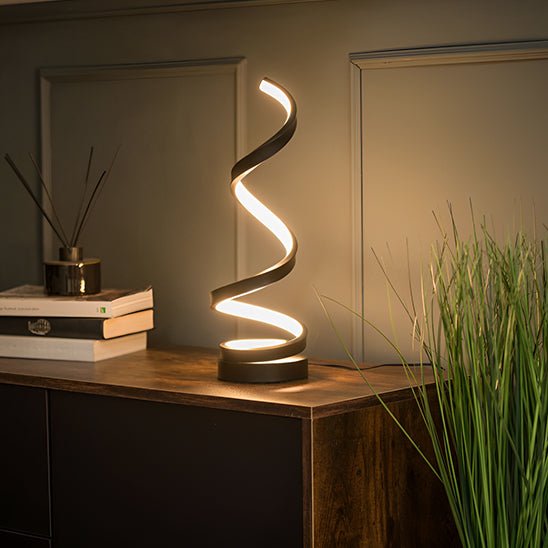 lampe de chevet spirale noire allumée sur une crédence en bois foncé un diffuseur de parfum à bâtonnets en bois est sur des livres des feuilles vertes sur le sol un mur couleur beige derrière un style nordique chaud en soirée