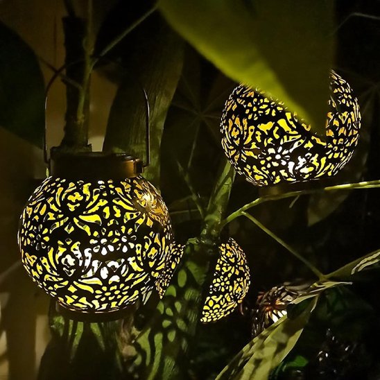 Lanterne solaire Sultana boule métal ajouré motifs organiques deux sont suspendues dans feuillage tropical nuit, lumière dorée chaude