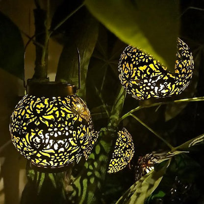 Lanterne solaire Sultana boule métal ajouré motifs organiques deux sont suspendues dans feuillage tropical nuit, lumière dorée chaude
