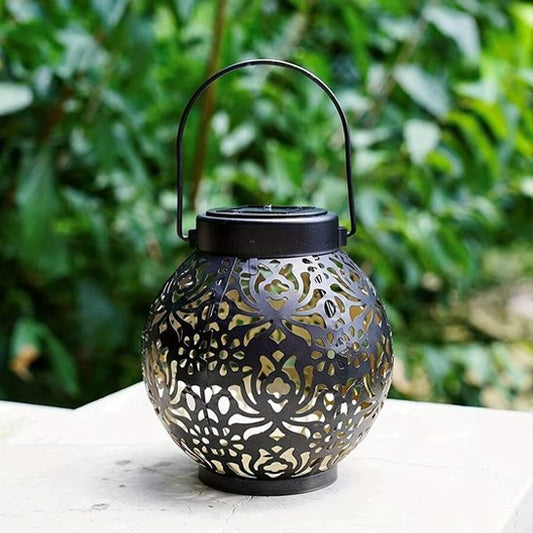 Lanterne solaire boule noire métal ajouré motifs orientaux posée sur table blanche, fond végétal vert, décoration jardin terrasse