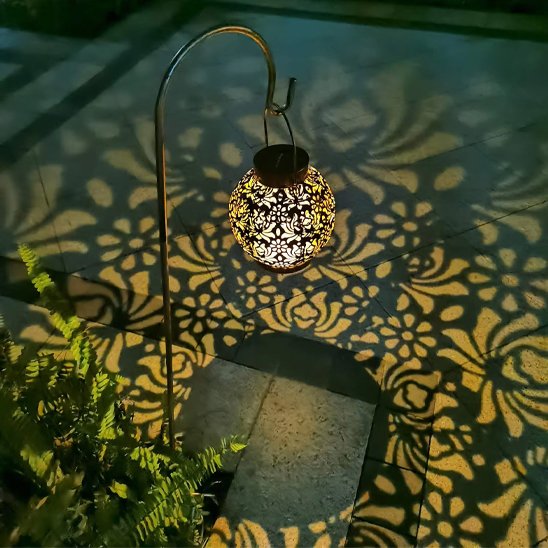 Lanterne solaire boule métal ajouré suspendue sur piquet arqué projetant grands motifs floraux dorés au sol terrasse nuit
