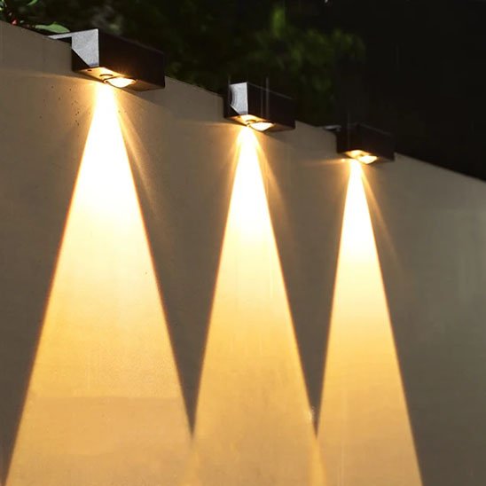 Luminaire extérieur trois noires alignées sur clôture extérieure diffusant lumière chaude triangulaire vers le bas